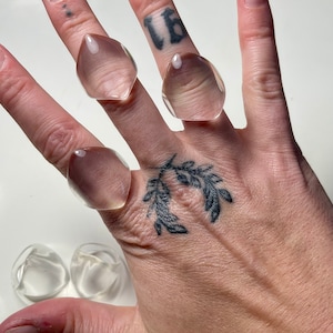 May include: Clear, geometric resin rings are displayed on a hand. The rings have a faceted design and a translucent appearance. The rings are a light, neutral color and appear to be handmade. The rings are a unique fashion accessory.