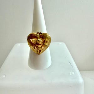 May include: A gold-colored ring with a heart-shaped design. The ring features an embossed image of a cherub with wings. The ring is displayed on a white cone-shaped ring holder.