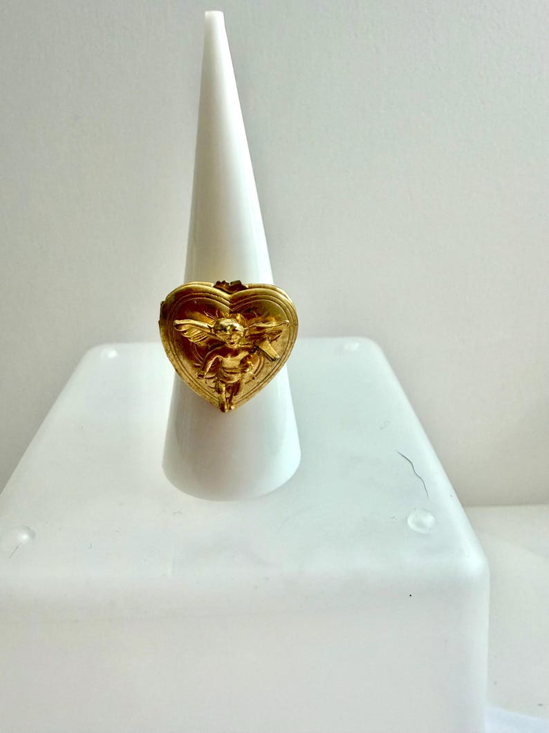 May include: A gold-colored ring with a heart-shaped design. The ring features an embossed image of a cherub with wings. The ring is displayed on a white cone-shaped ring holder.