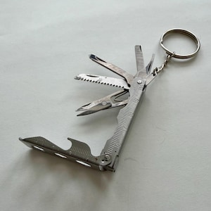 Op de afbeelding: Een zilveren multi-tool sleutelhanger met diverse uitgevouwen gereedschappen, waaronder een zaag, mes en flesopener. De tool heeft een liniaal aan de zijkant en een sleutelhanger. De multi-tool is ontworpen voor dagelijks gebruik en gemak.