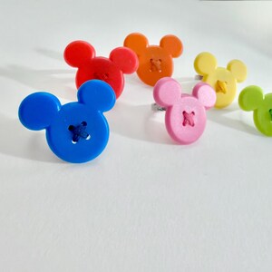 May include: Six colourful rings, each featuring a button shaped like a mouse head. The rings are in shades of blue, red, orange, pink, yellow, and green. The buttons have stitched details.