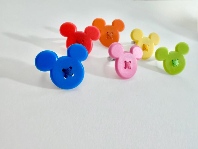 May include: Six colourful rings, each featuring a button shaped like a mouse head. The rings are in shades of blue, red, orange, pink, yellow, and green. The buttons have stitched details.