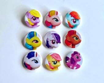 Può includere: Nove bottoni rotondi colorati con facce di pony dei cartoni animati. Ogni bottone presenta un design di pony diverso con colori variabili come rosa, giallo, blu e viola. I bottoni sono disposti in una griglia 3x3 su sfondo bianco.