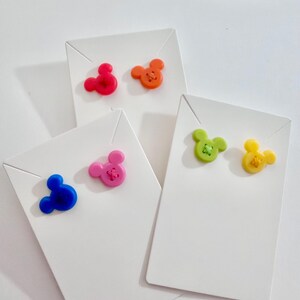 May include: Colorful stud earrings in the shape of a mouse head, presented on white display cards. The earrings come in red, orange, blue, pink, green, and yellow. The earrings have a button-like appearance.