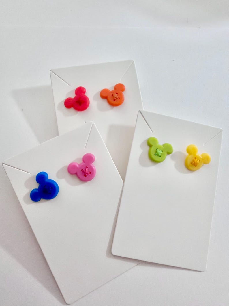 May include: Colorful stud earrings in the shape of a mouse head, presented on white display cards. The earrings come in red, orange, blue, pink, green, and yellow. The earrings have a button-like appearance.