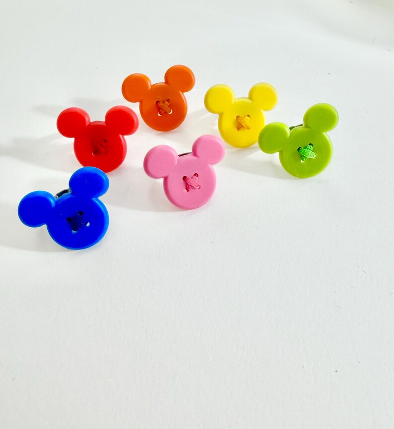 May include: Six colourful buttons in the shape of a mouse's head. The buttons are in red, orange, yellow, green, pink, and blue. Each button has a contrasting thread sewn through the centre.