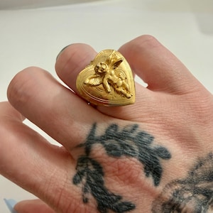 May include: A gold-colored ring with a heart-shaped face. The ring features a detailed relief of a cherub on the heart. The ring is worn on a finger. The ring is a piece of jewelry.