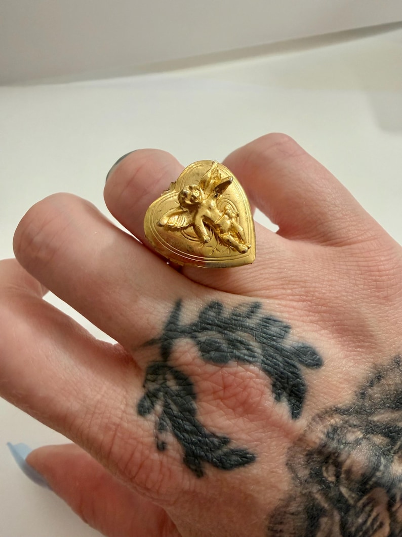 May include: A gold-colored ring with a heart-shaped face. The ring features a detailed relief of a cherub on the heart. The ring is worn on a finger. The ring is a piece of jewelry.