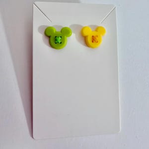May include: A pair of stud earrings in the shape of Mickey Mouse. One earring is lime green with green stitching, and the other is yellow with orange stitching. The earrings are displayed on a white earring card.
