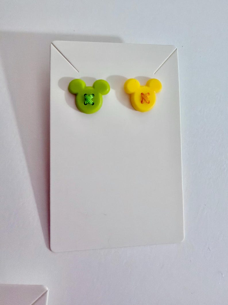May include: A pair of stud earrings in the shape of Mickey Mouse. One earring is lime green with green stitching, and the other is yellow with orange stitching. The earrings are displayed on a white earring card.
