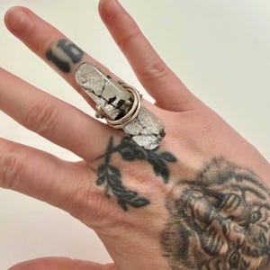 May include: A ring featuring a long, rectangular piece of silver-toned dichroic glass with a cracked, reflective surface texture. The glass is secured to the finger by a coiled silver-colored metal wire band that wraps around the center.