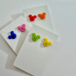 May include: A collection of white cardstock tags, each adorned with a colorful, cartoon mouse-shaped button. The buttons are in various colors: red, orange, pink, blue, green, and yellow. The tags are arranged in a slightly overlapping pattern.