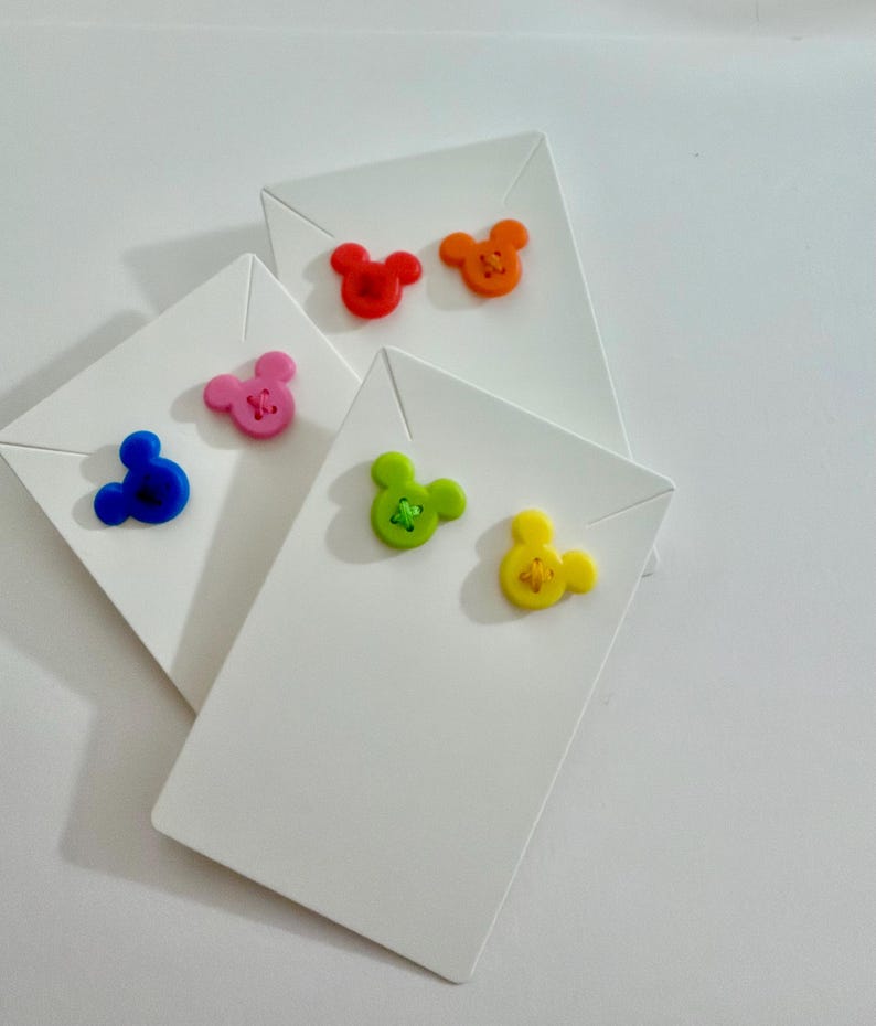 May include: A collection of white cardstock tags, each adorned with a colorful, cartoon mouse-shaped button. The buttons are in various colors: red, orange, pink, blue, green, and yellow. The tags are arranged in a slightly overlapping pattern.