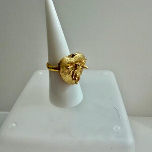 May include: A gold-colored ring with a heart-shaped design. The ring features intricate detailing on the heart, with a small bow and other embellishments. The ring is displayed on a white cone-shaped stand against a white background.