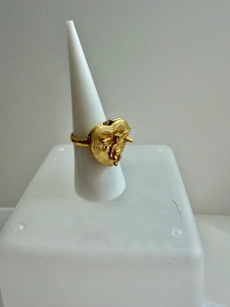 May include: A gold-colored ring with a heart-shaped design. The ring features intricate detailing on the heart, with a small bow and other embellishments. The ring is displayed on a white cone-shaped stand against a white background.