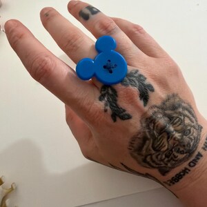 May include: A bright blue Mickey Mouse-shaped ring made from a large button. The ring features a cross-stitch detail in the centre, and the design includes two smaller circles for the ears. A fun and playful accessory.