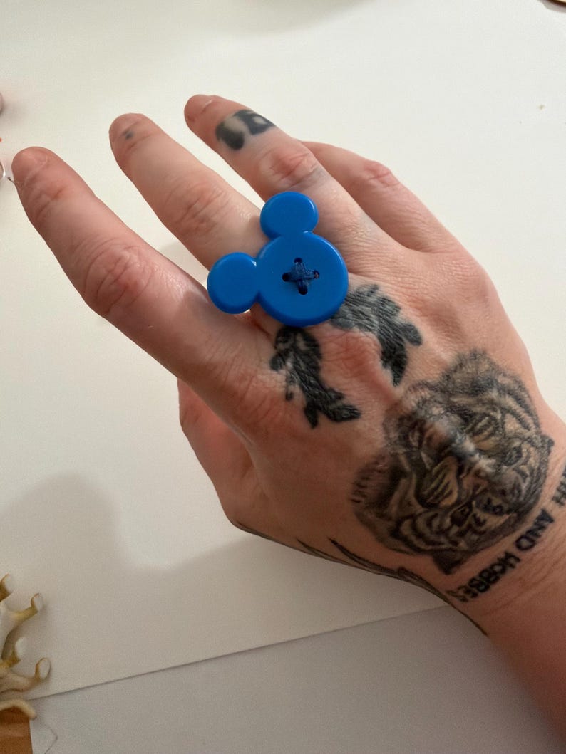 May include: A bright blue Mickey Mouse-shaped ring made from a large button. The ring features a cross-stitch detail in the centre, and the design includes two smaller circles for the ears. A fun and playful accessory.