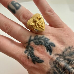 May include: A gold-colored ring in the shape of a heart, adorned with a detailed relief of cherubs. The ring is worn on a finger. The ring's design is intricate, with the cherubs appearing to be in flight. The ring is a statement piece.