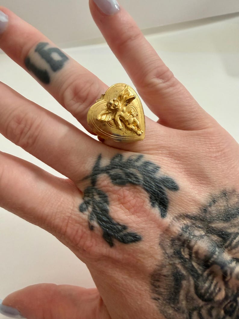 May include: A gold-colored ring in the shape of a heart, adorned with a detailed relief of cherubs. The ring is worn on a finger. The ring's design is intricate, with the cherubs appearing to be in flight. The ring is a statement piece.