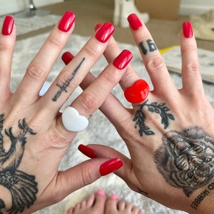 May include: Two hands with red nail polish, each wearing a heart-shaped ring. One ring is white, the other is red. The hands have tattoos of a dagger, a tiger, and other designs.