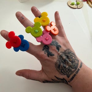 May include: A hand shows six colourful, Mickey Mouse-shaped buttons. The buttons are red, blue, yellow, green, pink, and orange. Each button has a contrasting colour thread sewn through it. The hand has a tiger tattoo and the words "CALVIN AND HOBBES".