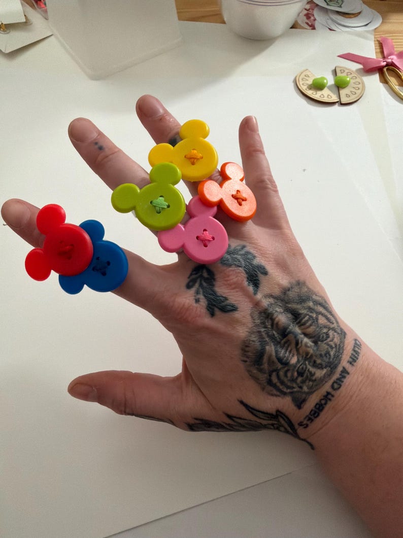 May include: A hand shows six colourful, Mickey Mouse-shaped buttons. The buttons are red, blue, yellow, green, pink, and orange. Each button has a contrasting colour thread sewn through it. The hand has a tiger tattoo and the words "CALVIN AND HOBBES".