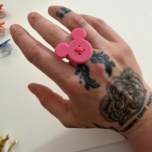 May include: A bright pink button ring in the shape of Mickey Mouse. The ring has a large, round button with two smaller circles for ears, and a cross-stitch detail in the centre. A playful, novelty accessory.