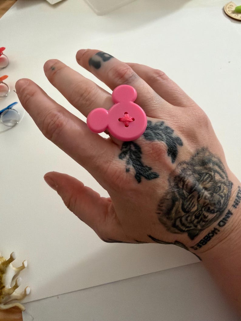 May include: A bright pink button ring in the shape of Mickey Mouse. The ring has a large, round button with two smaller circles for ears, and a cross-stitch detail in the centre. A playful, novelty accessory.
