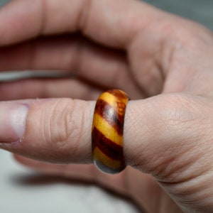 May include: A wooden ring with a striped pattern of light brown and dark brown. The ring is worn on a finger, showcasing the natural wood grain and polished finish. The ring's design features a smooth, rounded shape.