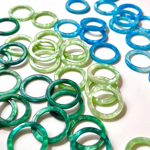 Puede incluir: Una colección de anillos de resina translúcida en varios tonos de verde y azul. Los anillos están dispersos sobre una superficie blanca, mostrando una gama de colores que van desde el esmeralda oscuro hasta el aguamarina claro. Los anillos miden aproximadamente 2,5 cm de diámetro.