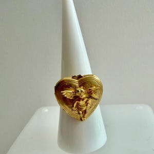 May include: A gold-colored ring with a heart-shaped locket design. The locket features a detailed relief of a cherub with wings. The ring is displayed on a white, cone-shaped stand, showcasing its intricate details and design.