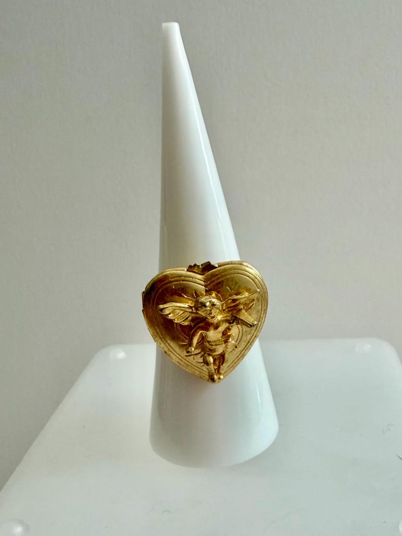 May include: A gold-colored ring with a heart-shaped locket design. The locket features a detailed relief of a cherub with wings. The ring is displayed on a white, cone-shaped stand, showcasing its intricate details and design.