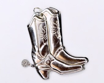 Puede incluir: Colgante de botas de vaquero plateadas con intrincados detalles florales y una espuela. El colgante tiene un lazo para colgar y probablemente está hecho de metal. Las botas son un diseño clásico del Oeste.