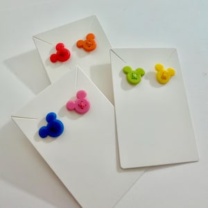 May include: A collection of colorful Mickey Mouse-shaped earrings displayed on white earring cards. The earrings are in various colors, including red, orange, pink, blue, green, and yellow. Each earring has a stitched detail. The cards are arranged at different angles.
