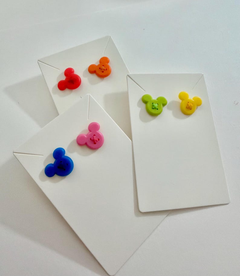 May include: A collection of colorful Mickey Mouse-shaped earrings displayed on white earring cards. The earrings are in various colors, including red, orange, pink, blue, green, and yellow. Each earring has a stitched detail. The cards are arranged at different angles.