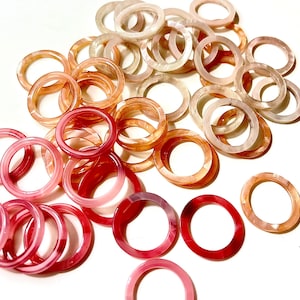 Puede incluir: Una colección de anillos translúcidos en tonos rosa, rojo y melocotón. Los anillos están dispersos sobre una superficie blanca, mostrando sus diferentes tonos y acabado brillante. Los anillos parecen estar hechos de resina o material acrílico.