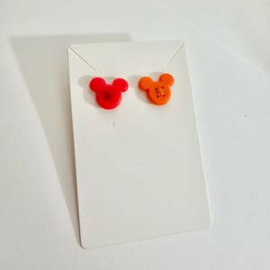 May include: A pair of stud earrings in the shape of a mouse head. One earring is red, the other is orange. The earrings are displayed on a white earring card.
