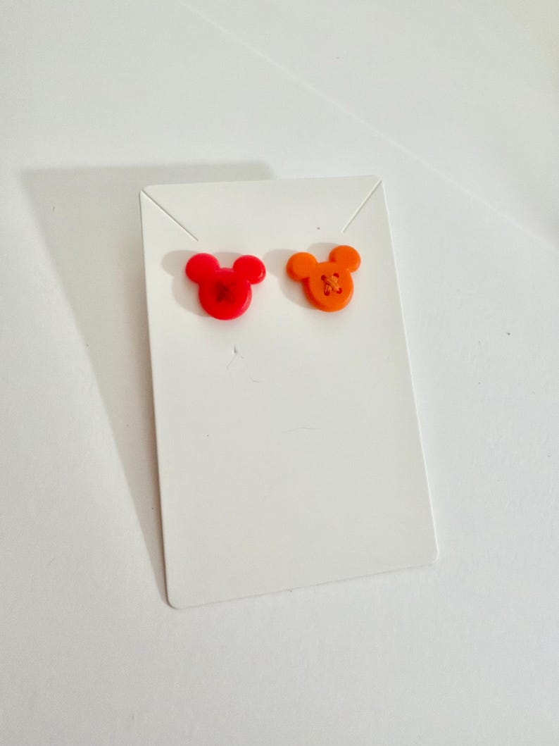 May include: A pair of stud earrings in the shape of a mouse head. One earring is red, the other is orange. The earrings are displayed on a white earring card.