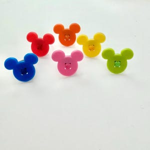 May include: Seven colourful buttons in the shape of a mouse's head. The buttons are in shades of red, orange, yellow, blue, pink, and green. Each button has a stitched detail in the centre.