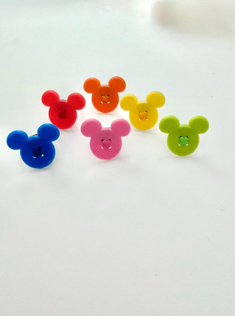 May include: Seven colourful buttons in the shape of a mouse's head. The buttons are in shades of red, orange, yellow, blue, pink, and green. Each button has a stitched detail in the centre.
