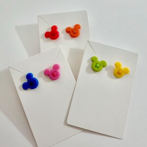 May include: A collection of colorful stud earrings shaped like a mouse head. The earrings are red, orange, pink, blue, green, and yellow. Each pair is displayed on a white earring card.