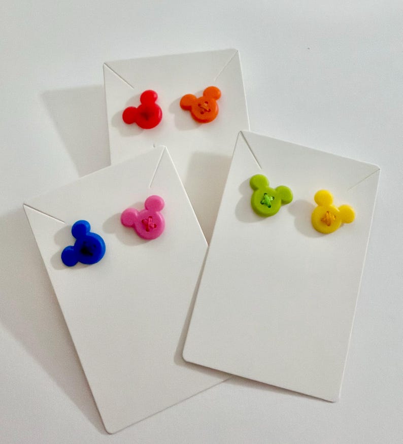 May include: A collection of colorful stud earrings shaped like a mouse head. The earrings are red, orange, pink, blue, green, and yellow. Each pair is displayed on a white earring card.