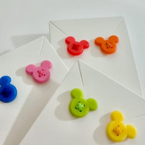 May include: A collection of white folded cards, each adorned with a colorful Mickey Mouse-shaped button. Buttons are in shades of red, orange, pink, blue, green, and yellow. The buttons have a cross-stitch design in the center.