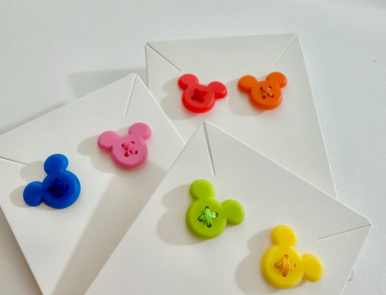 May include: A collection of white folded cards, each adorned with a colorful Mickey Mouse-shaped button. Buttons are in shades of red, orange, pink, blue, green, and yellow. The buttons have a cross-stitch design in the center.