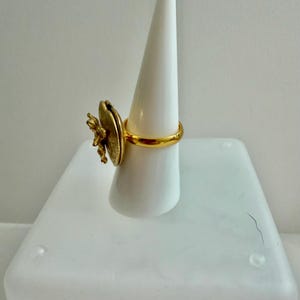 May include: A gold-colored ring with a large, textured, circular design and a smaller, detailed floral accent. The ring is displayed on a white, conical stand, showcasing its intricate details and design. The ring's band is a simple, polished gold.