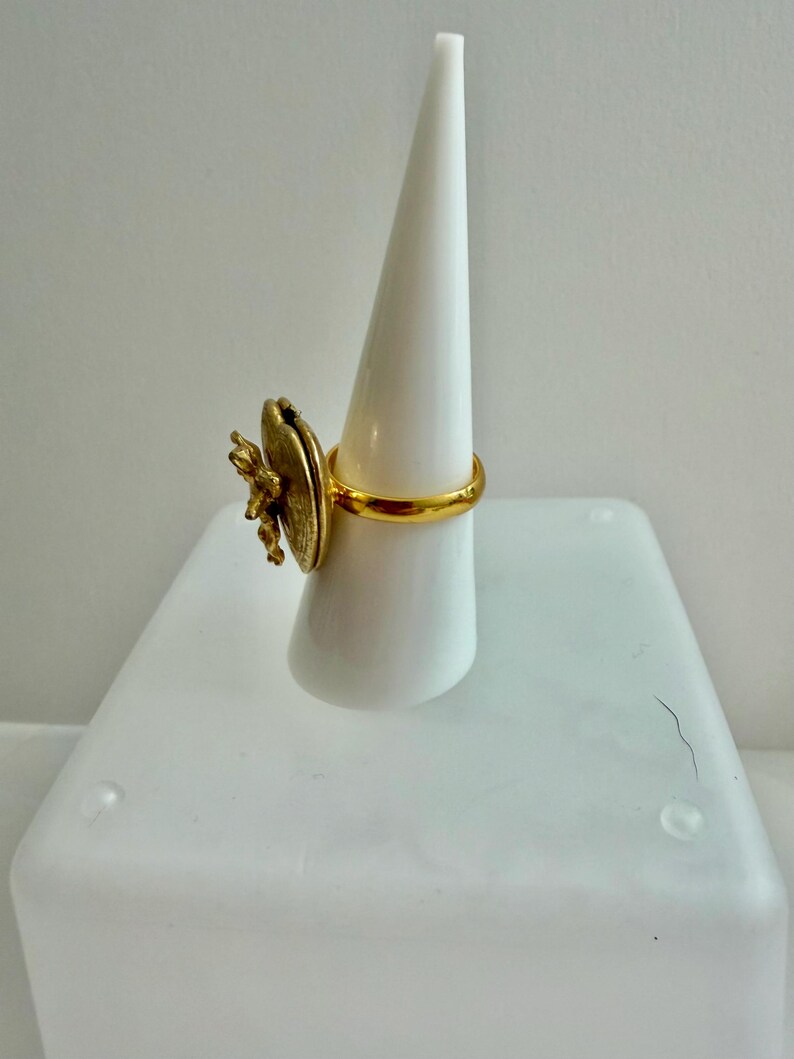 May include: A gold-colored ring with a large, textured, circular design and a smaller, detailed floral accent. The ring is displayed on a white, conical stand, showcasing its intricate details and design. The ring's band is a simple, polished gold.