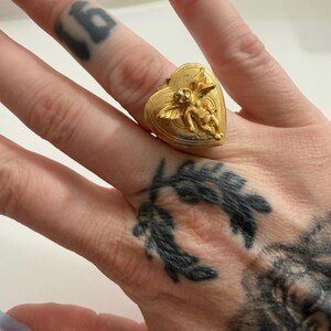May include: A gold-colored ring in the shape of a heart, featuring a detailed relief of two cherubs. The ring is worn on a finger, showcasing its intricate design and vintage aesthetic. The ring's heart shape adds a romantic touch.