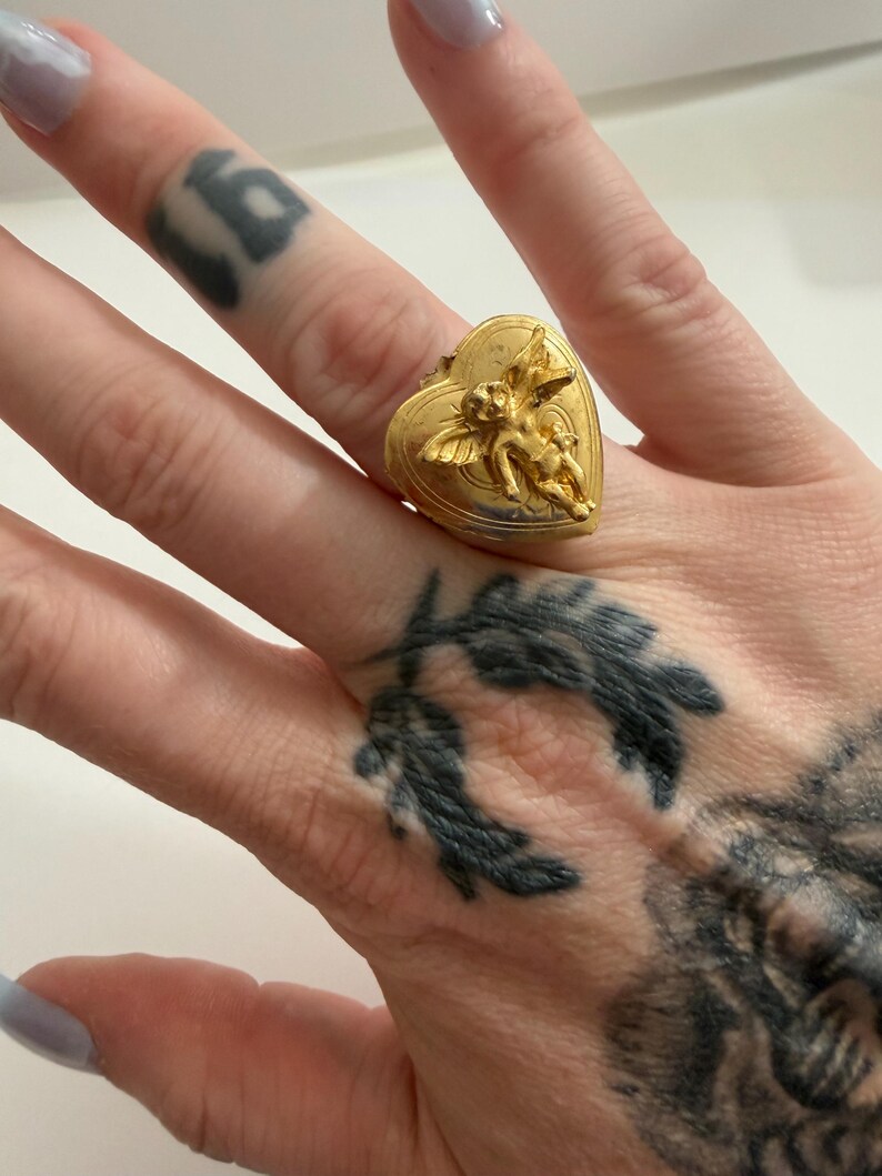 May include: A gold-colored ring in the shape of a heart, featuring a detailed relief of two cherubs. The ring is worn on a finger, showcasing its intricate design and vintage aesthetic. The ring's heart shape adds a romantic touch.