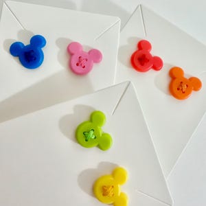May include: A collection of colorful, Mickey Mouse-shaped buttons in blue, pink, red, orange, green, and yellow. Each button is sewn onto a white square. The buttons are arranged on a white surface.