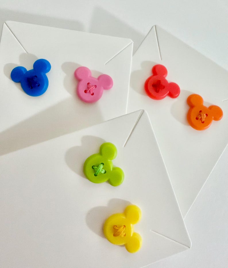 May include: A collection of colorful, Mickey Mouse-shaped buttons in blue, pink, red, orange, green, and yellow. Each button is sewn onto a white square. The buttons are arranged on a white surface.